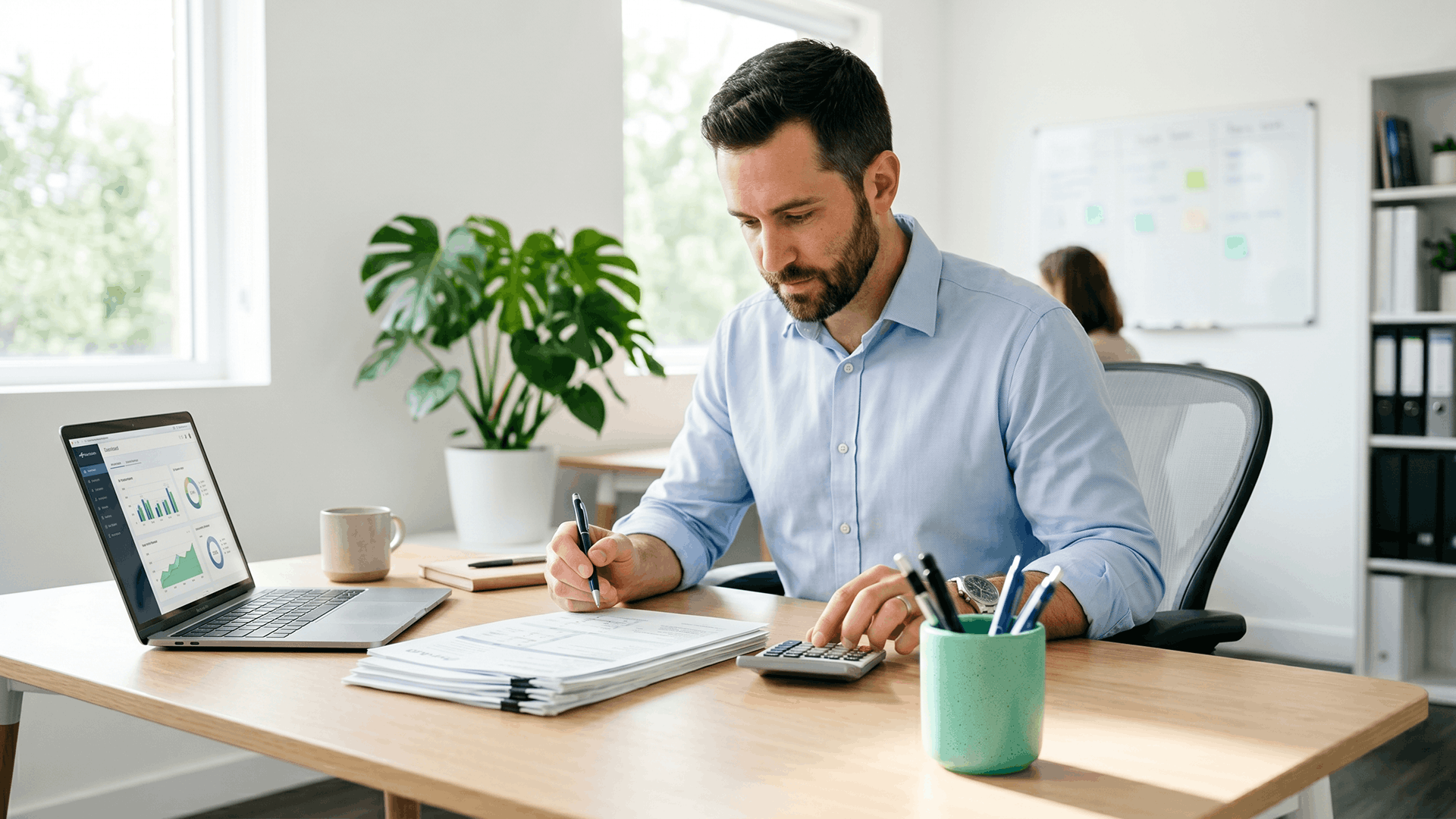 Practice finance leader at a wooden desk working through a stack of prior authorization paperwork with a calculator beside an open laptop showing a charts dashboard, calculating the fully loaded cost per request