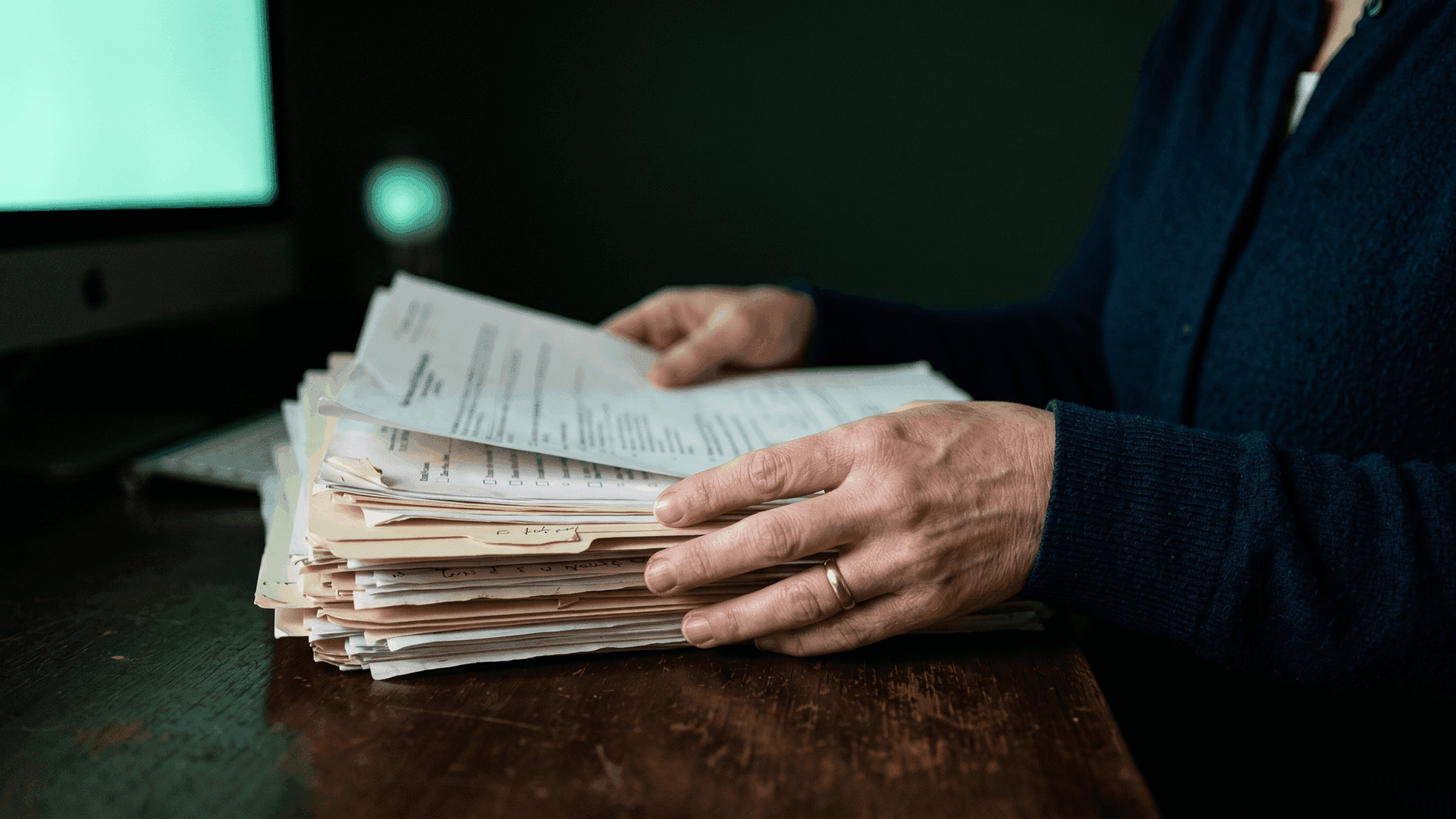 Hands sorting through a thick stack of prior authorization paperwork on a dark wooden desk, with a softly glowing mint-green monitor in the background — the manual paperwork burden FQHC coordinators face across four to eight Medicaid MCO portals