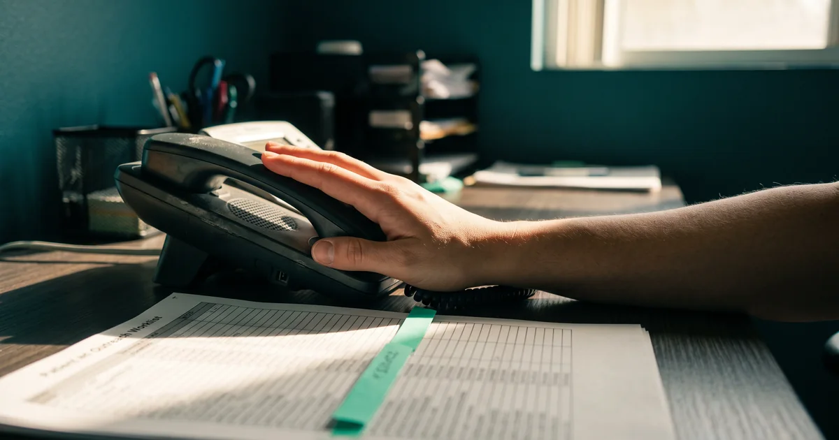 Outreach worklist beside the desk phone A hand resting on a desk phone handset beside a printed patient outreach worklist with a mint tab in a sunlit office