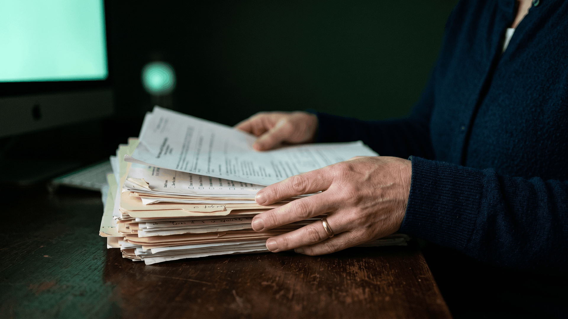 Hands sorting through a thick stack of prior authorization paperwork on a dark wooden desk, with a softly glowing mint-green monitor in the background — the manual paperwork burden FQHC coordinators face across four to eight Medicaid MCO portals
