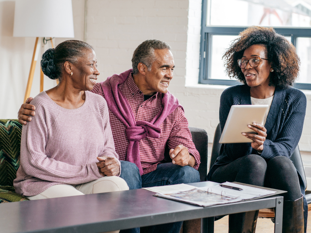 Two patients sit on a couch and smile while looking at a document held by a healthcare professional